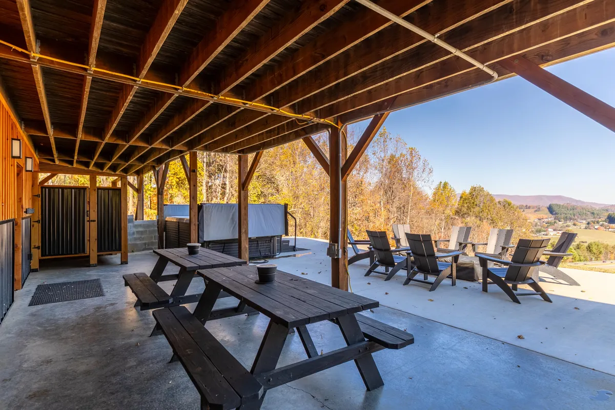 This image shows a spacious outdoor deck featuring wooden picnic tables, reclining chairs, a bubbling hot tub, and surrounded by natural greenery. The setup encourages group dining, relaxation, and soaking in the hot tub while appreciating the natural views.