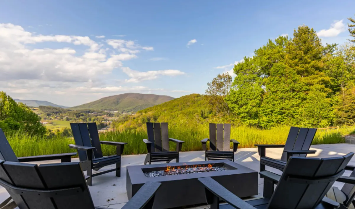 A cozy fire pit surrounded by Adirondack chairs sits atop a grassy knoll, with the Blue Ridge mountains providing a dramatic backdrop. The scene invites guests to gather outdoors, enjoy the sunset, and stargaze in a communal, natural setting.