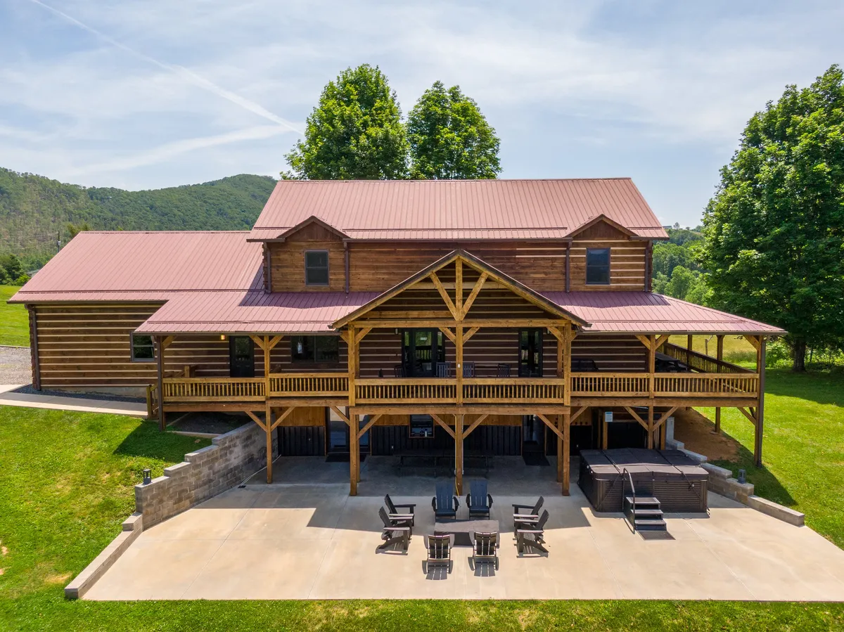This image features a log cabin with a large wraparound deck, outdoor seating areas, a bubbling hot tub, and the surrounding natural landscape. The deck is spacious, inviting, and clearly set up for gatherings and relaxation. The hot tub is visible, and the scene captures the essence of outdoor mountain living.