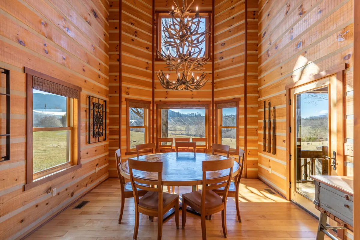 A round wooden dining table sits beneath a striking antler chandelier, surrounded by wooden walls and windows that allow in warm natural light. The setting feels intimate yet spacious, inviting guests to gather for meals and conversation.