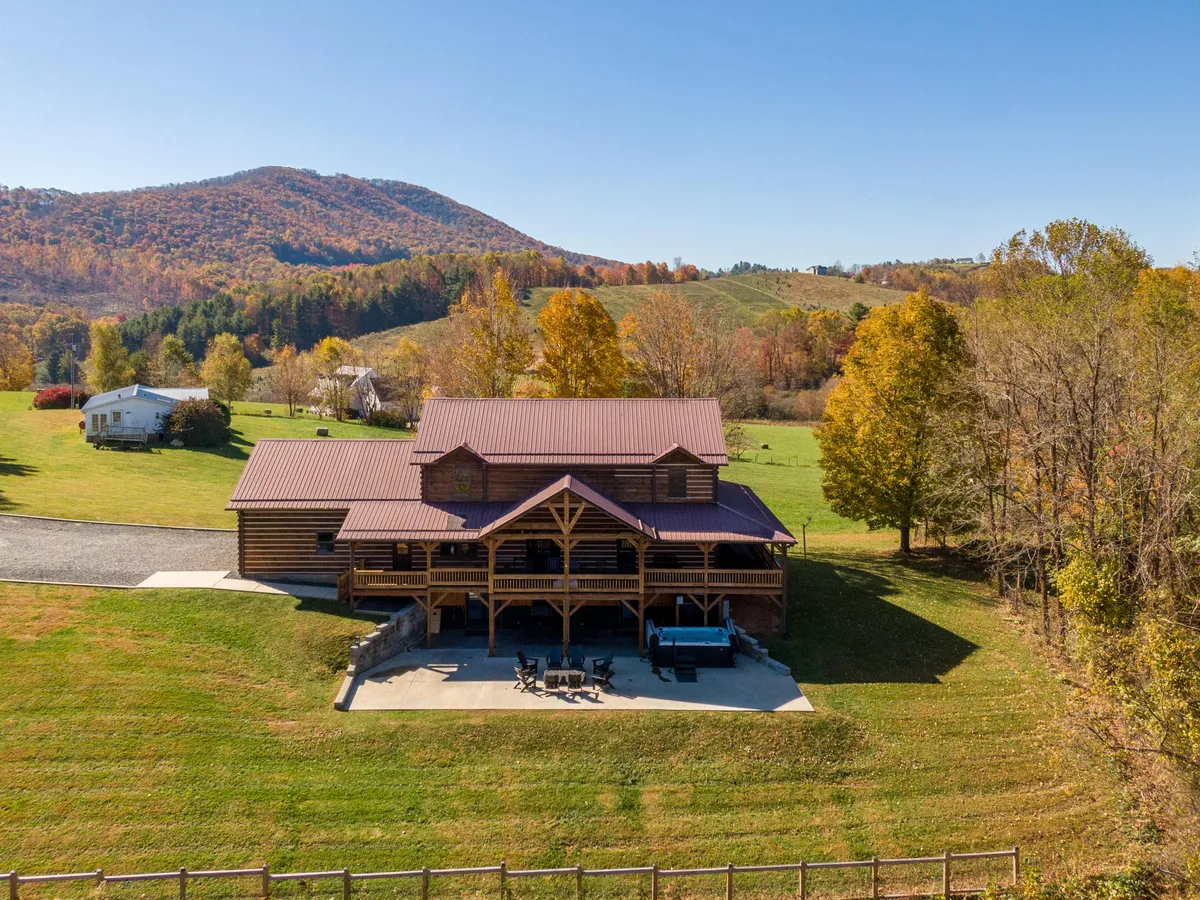 The log cabin sits surrounded by colorful autumn trees and majestic mountains, with a cozy outdoor fire pit area and a spacious porch. The setting showcases the mountain escape atmosphere and opportunities for outdoor gatherings.
