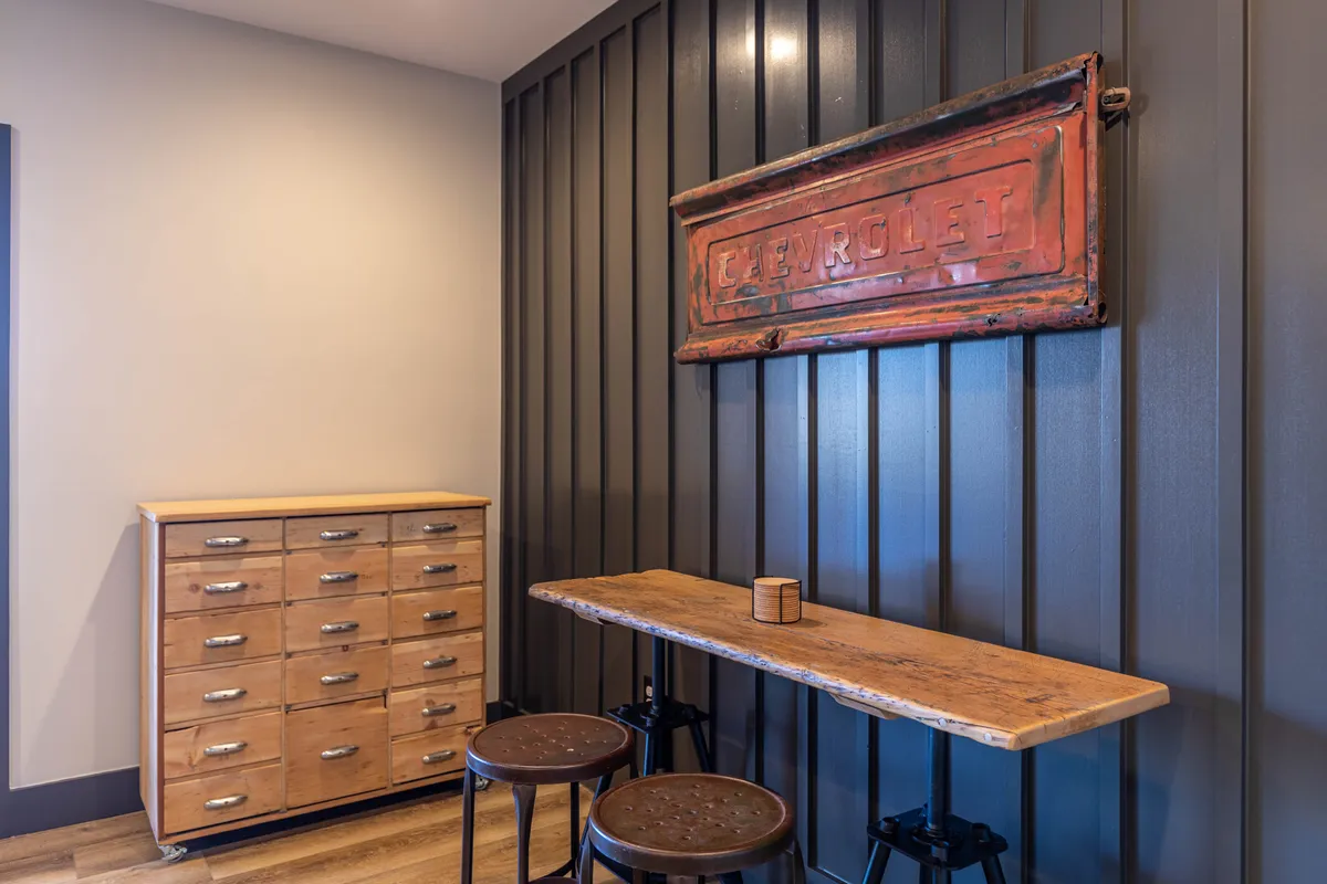 A stylish interior space with a vintage Chevrolet sign, a long wooden table, and metal stools. The nostalgic decor and comfortable setup create a perfect spot for social gatherings, coffee breaks, or casual chats before or after a game. The space feels inviting and encourages group interaction.