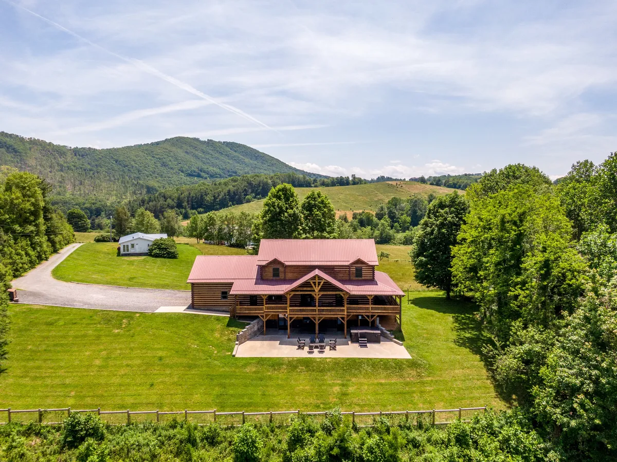 A classic log cabin is nestled amidst green lawns and surrounding mountains, with a patio equipped with comfortable chairs for gathering and relaxation. The landscape is inviting and highlights direct access to nature.
