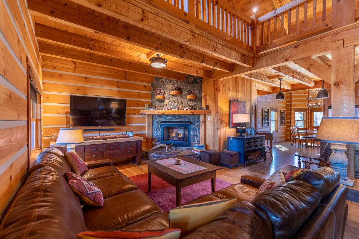 A spacious open-concept living area featuring a large leather sofa arranged around a prominent stone fireplace, with exposed wooden beams overhead. The TV is mounted beside the fireplace, and the dining area is visible in the background, emphasizing the room's seamless flow and communal design.