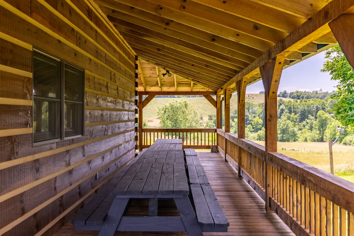 A wooden picnic table sits on a covered porch with sturdy beams, overlooking lush greenery in the background. The scene sets the stage for large family meals, sunrise coffees, or sunset celebrations, and captures the alfresco dining experience described in the section.