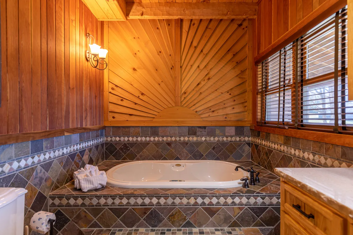 A luxurious bathroom featuring a jacuzzi tub, wooden wall with sunburst design, stone-tiled walls, and a window with blinds. The tub invites relaxation and is situated to take advantage of soft natural light, emphasizing comfort and self-care.