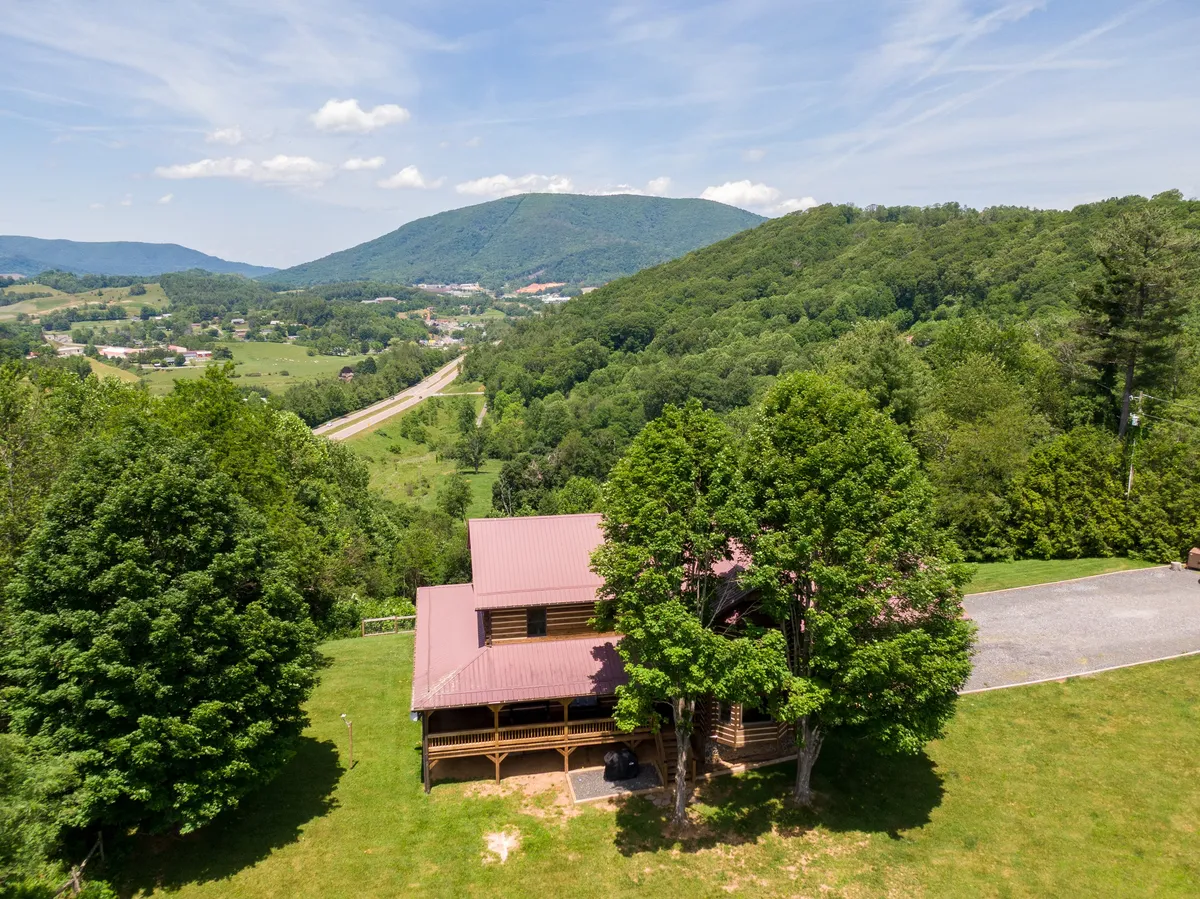 A sweeping exterior shot of the log cabin, surrounded by green lawns with a mountain backdrop. The image radiates a peaceful mood and shows the full context of the property—its setting, spacious lawn, and the mountains beyond.