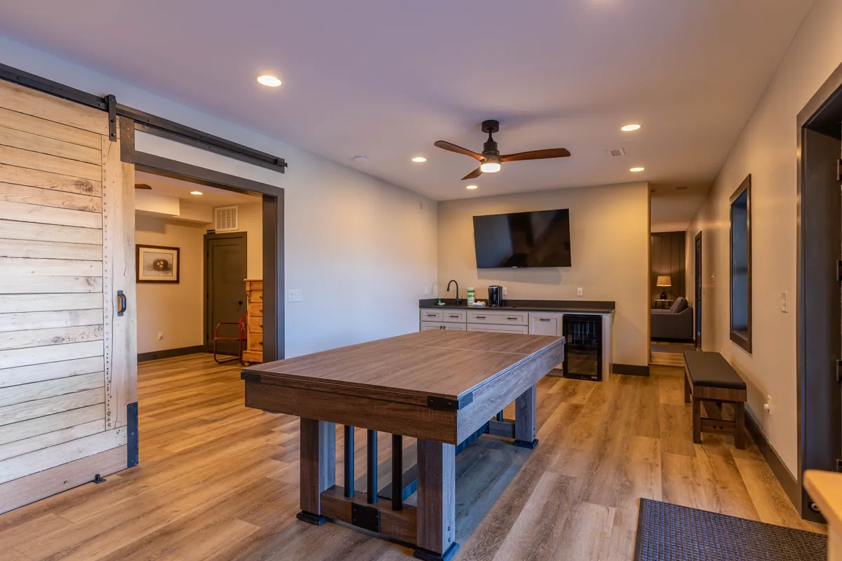 A spacious entertainment area featuring a large wooden table perfect for game nights, a sliding barn door, a flat-screen TV, and a kitchenette area. The room is set up for casual gatherings, games, and relaxing with friends or family, with plenty of seating and an inviting atmosphere.