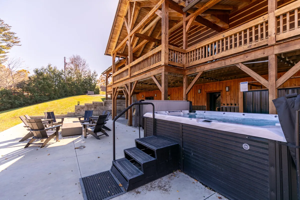 An inviting patio scene with a hot tub, outdoor seating area, and a wooden structure providing a covered space. The area is designed for socializing, relaxing in the hot tub, and enjoying the outdoors in comfort, regardless of weather.