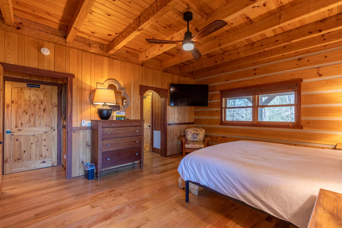 A welcoming bedroom with wooden ceiling beams, a dresser with a lamp, a bed with crisp white linens, a floral chair, and a television. The room feels both bright and inviting, ideal for families or guests seeking comfort and solitude.