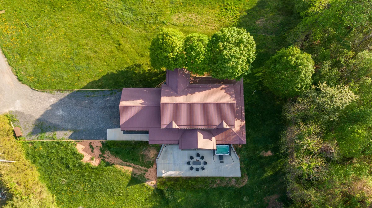 A wide view of the lodge exterior, showing the house with a red metal roof, lush greenery, an outdoor seating area with a custom fire pit, and a pool area. The space is ideal for outdoor gatherings, fire pit nights, and swimming under the open sky.